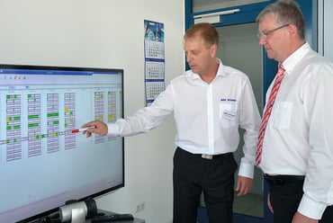 One man is explaining something to another, pointing to the monitor in front of them showing capacity planning.